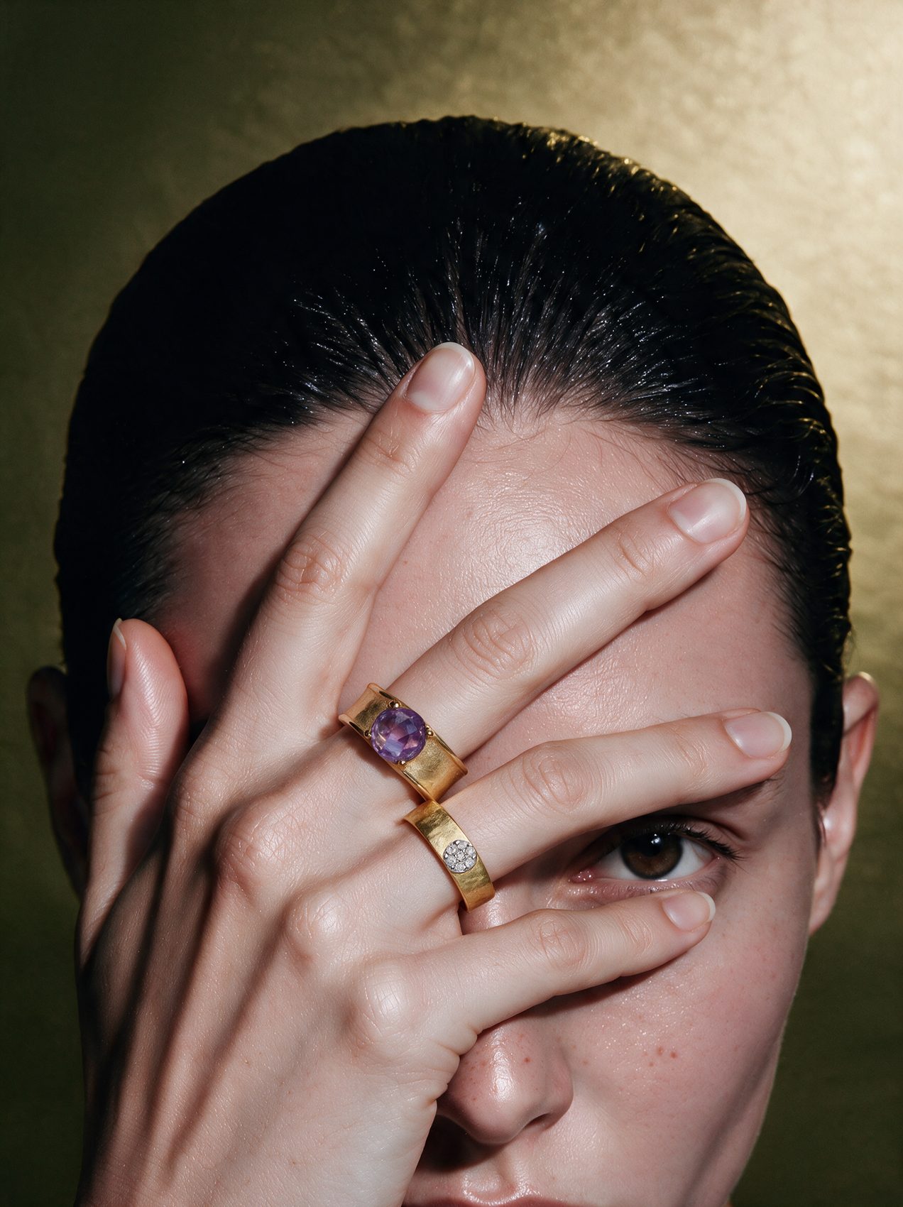 Hand across a face wearing a hammered gold band and an amethyst ring, against gold
